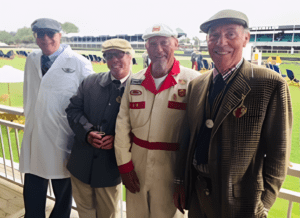 Four men in vintage-inspired outfits, including a lab coat and race suit, smile warmly at an outdoor event with green fields and grandstands.