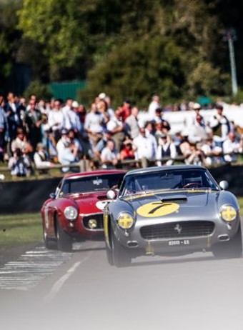 A silver vintage race car with yellow markings leads a red classic car