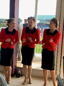 Three women in matching red sweaters and black skirts sing together
